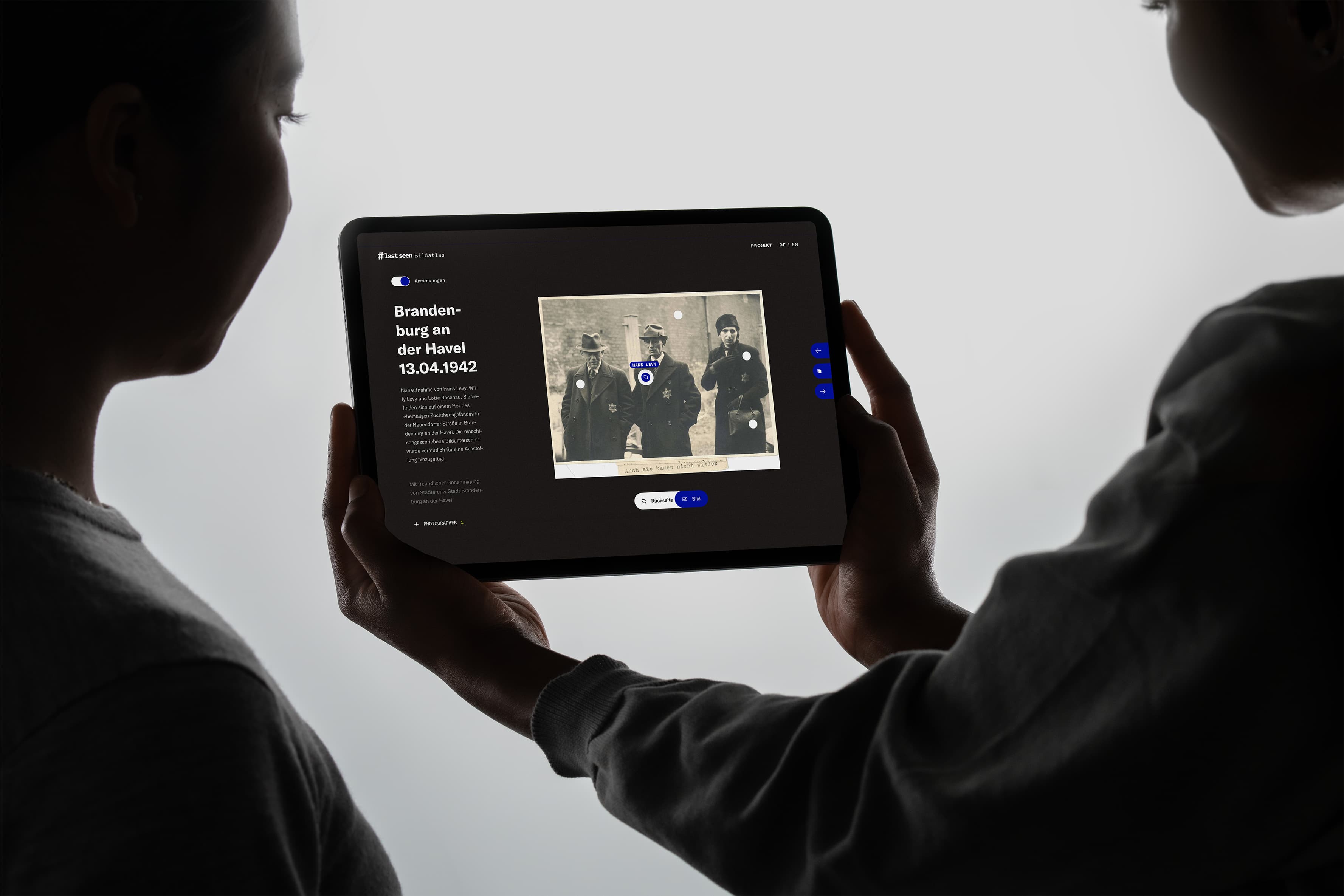 Two people holding a tablet displaying a sepia-toned photograph of soldiers. The screen shows annotations and text on the side. The surrounding area is softly lit, highlighting the device.