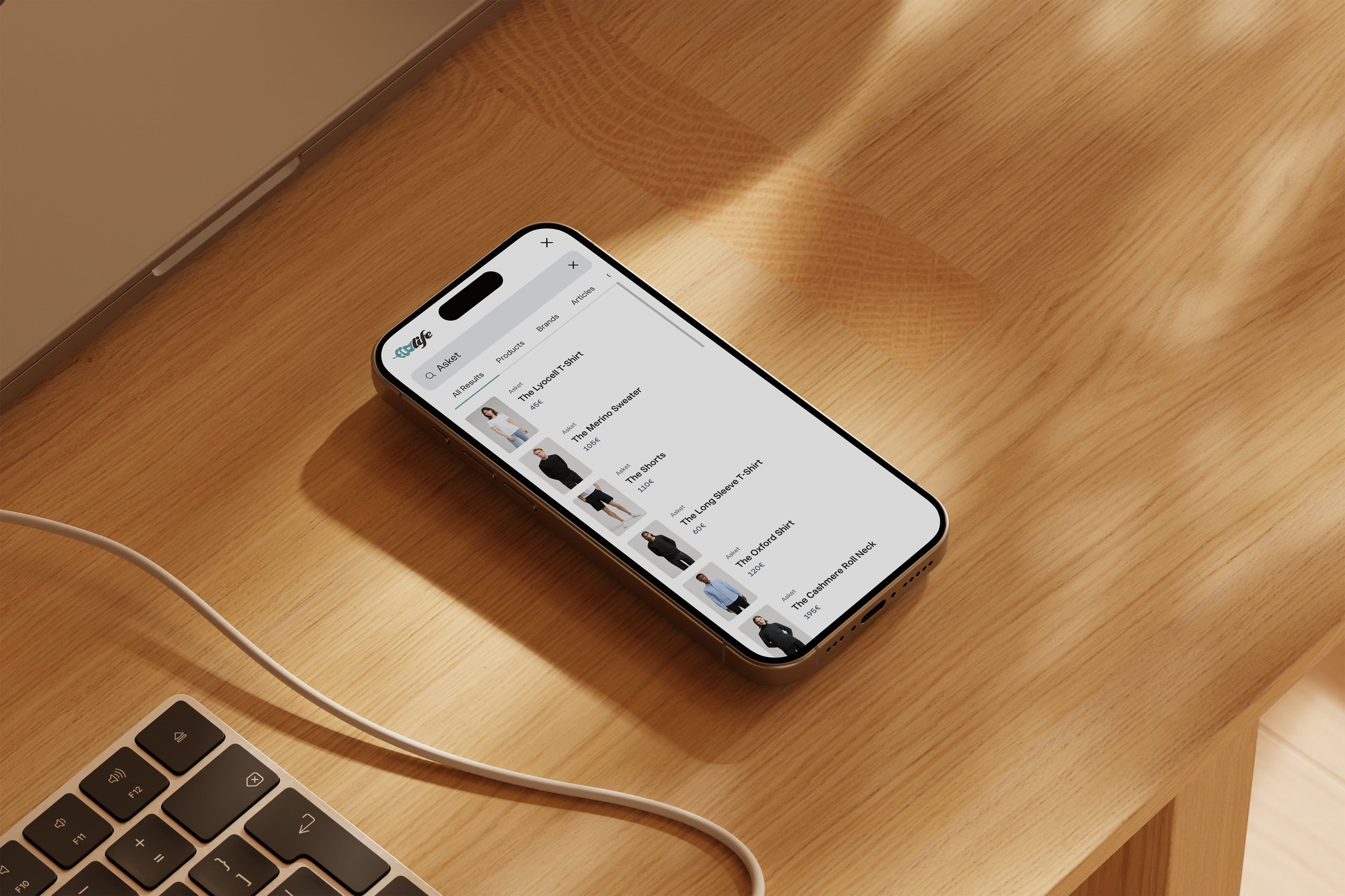A smartphone displaying a shopping app on a wooden desk next to an open laptop and a charging cable. The screen shows a list of clothing items available for purchase. Sunlight casts soft shadows on the surface.