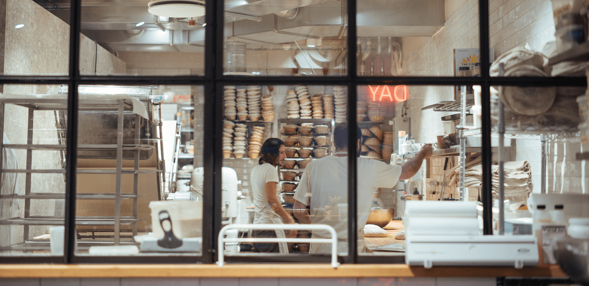 View through window into a bakery with two people, shelves full of bread and a workspace in the foreground