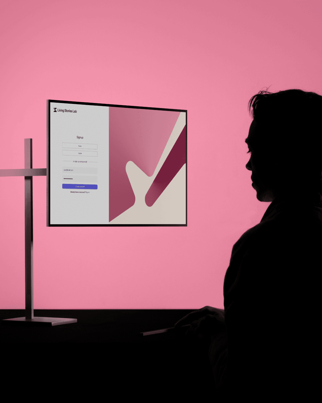 Silhouette of a person sitting at a desk, facing a monitor with a login screen displayed. The background is pink, and the screen shows a stylized abstract design.